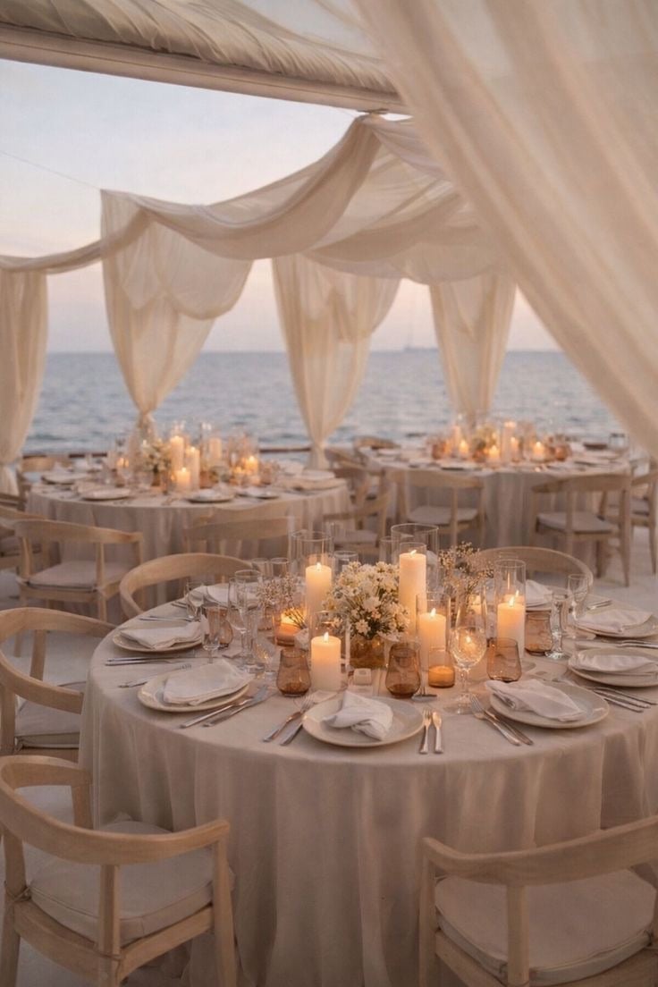 Elegant beachfront wedding reception setup with candlelit tables, sheer curtains, and ocean view at sunset