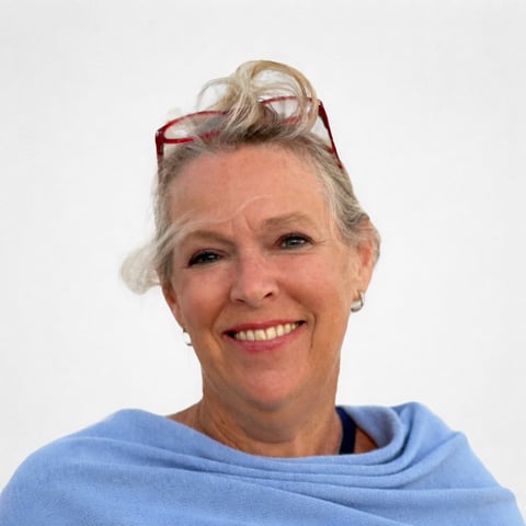 Portrait of a smiling middle-aged woman with blonde hair in a bun, red sunglasses on her head, wearing a blue sweater against a white background
