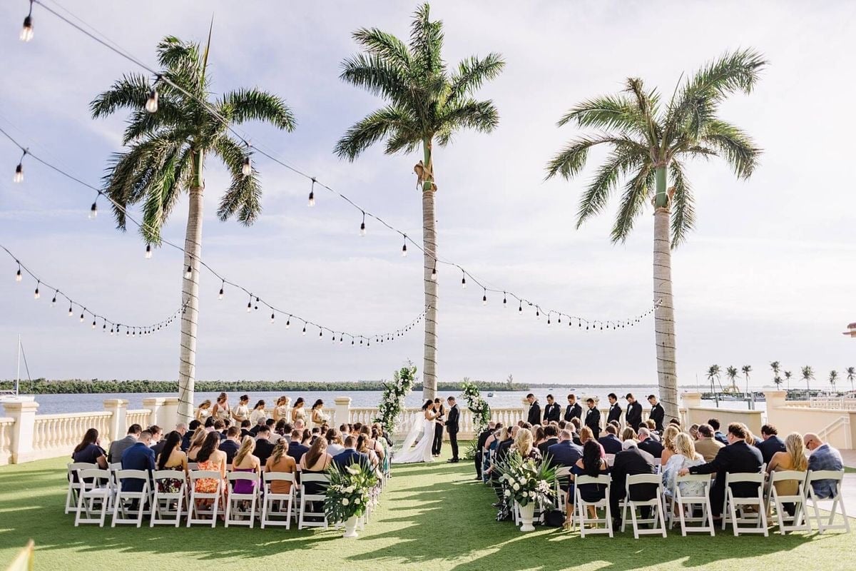 Outdoor wedding ceremony setup with guests seated in white chairs under three tall palm trees with string lights overhead and waterfront in background