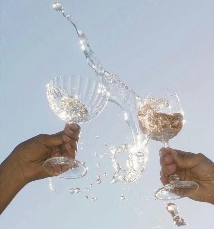 Two hands clinking champagne glasses with wine splashing against a clear blue sky