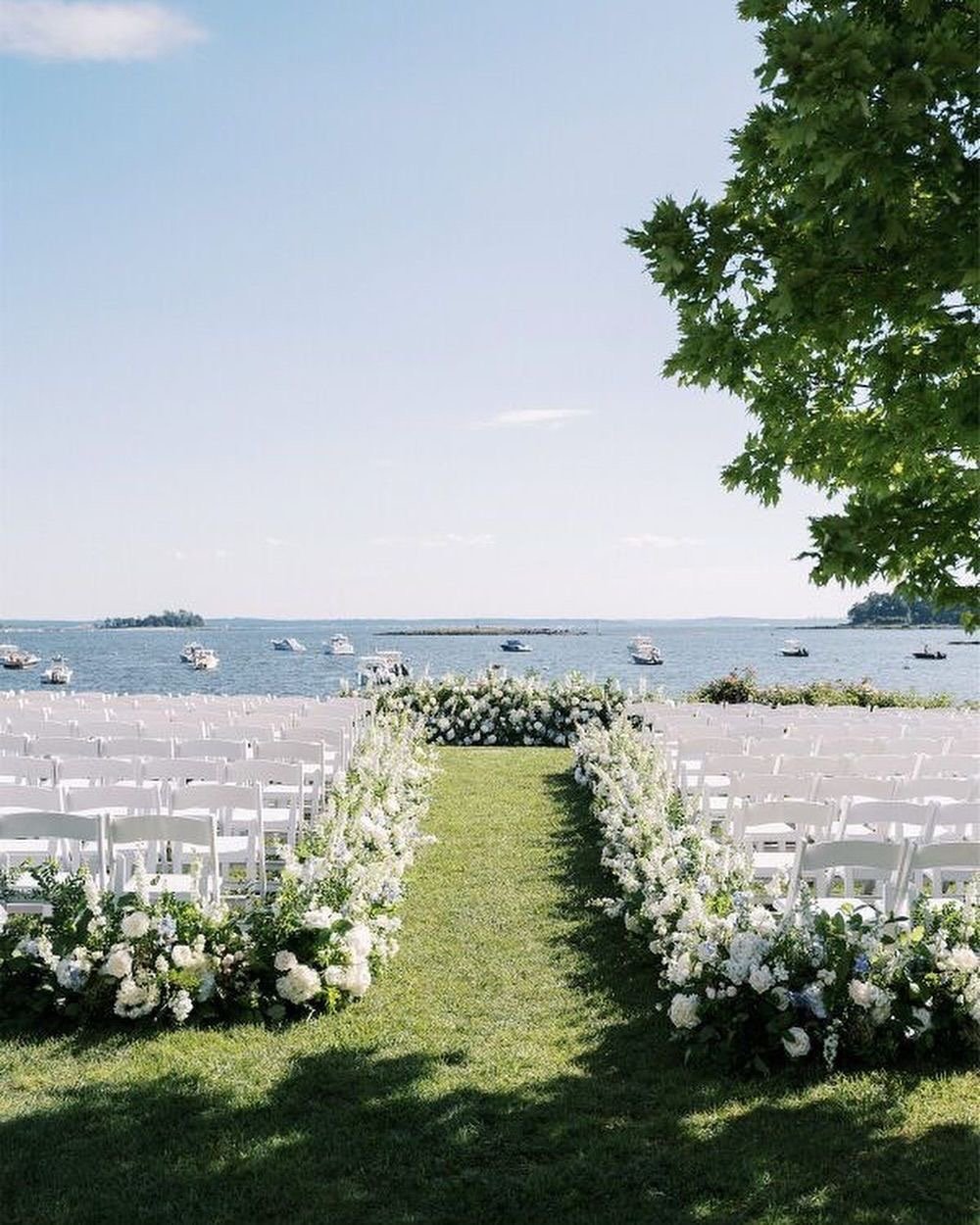 Waterfront wedding ceremony setup with white chairs lining a green aisle flanked by white floral arrangements and boats visible on the water