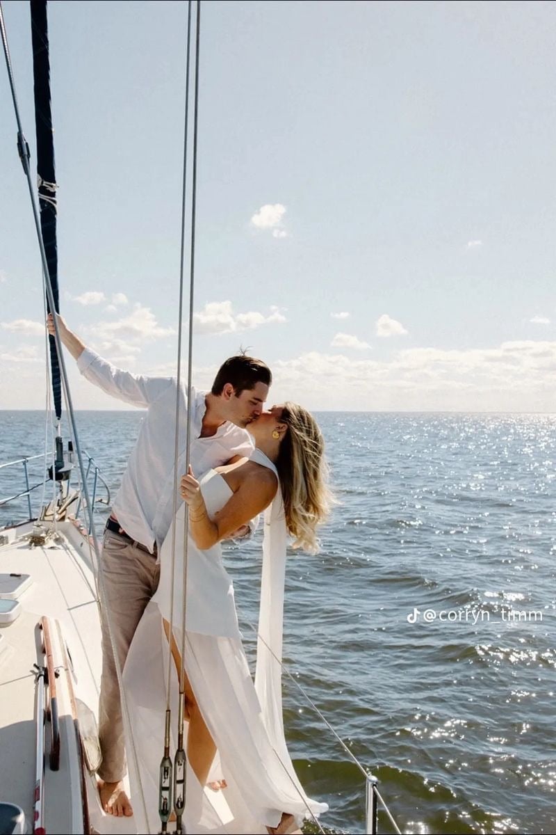 Couple in white clothing embracing on a sailboat with ocean and mountains in the background