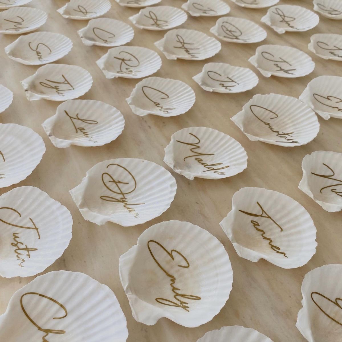 White scallop shells arranged on a neutral surface, each hand-inscribed with gold calligraphy names
